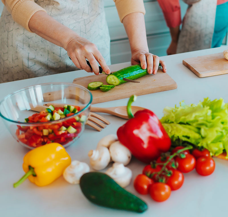 Close up image of female hands with knife cutting fresh cucumber on the table on domestic kitchen. Many ripe, tasty and colorful vegetables with olive oil. Vegan meal, healthy nutrition